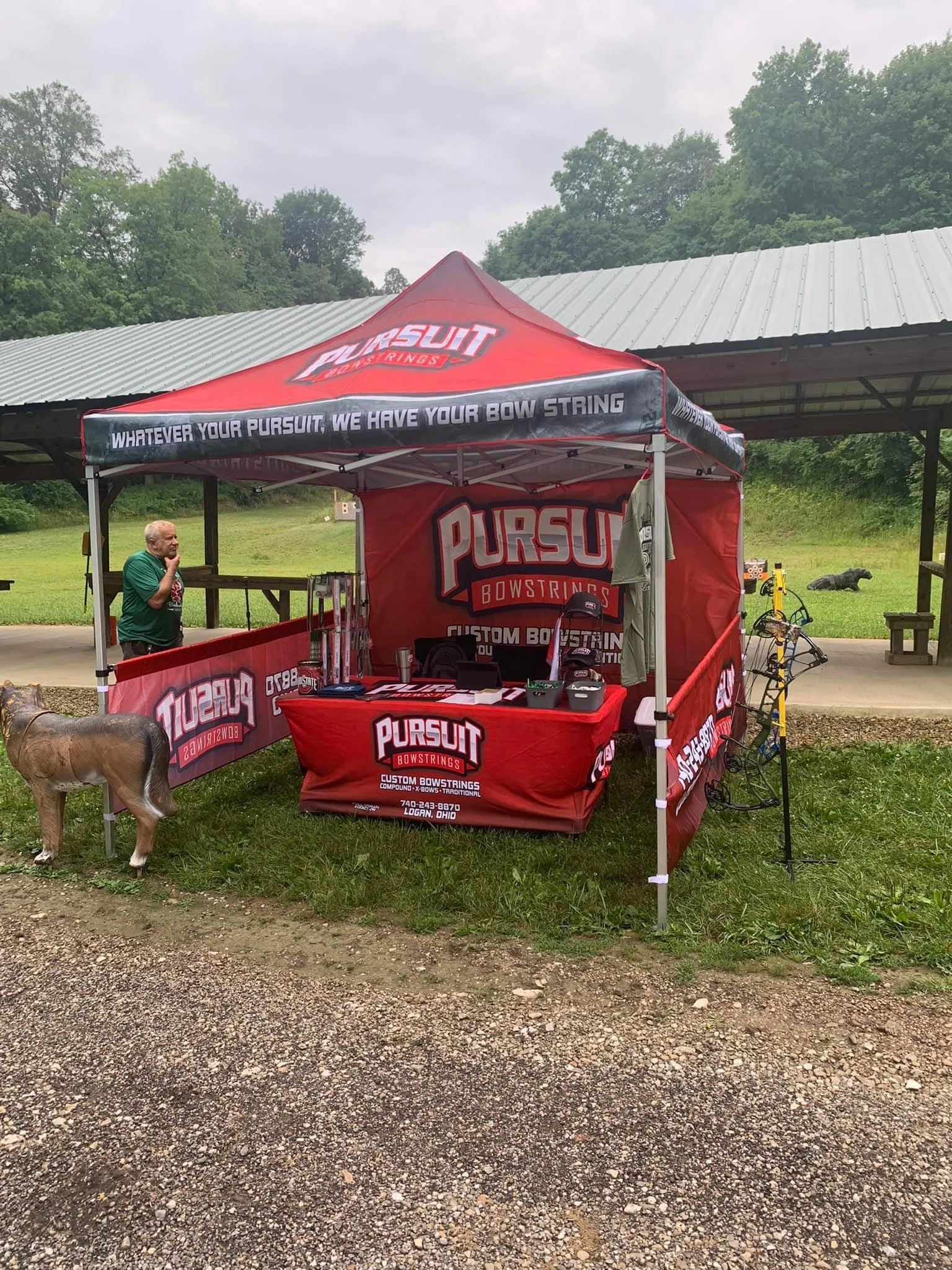 A Pursuit Bowstrings booth set up outdoors under a red canopy with tables and banners, with a man in a green shirt and a dog sculpture nearby, and trees in the background.