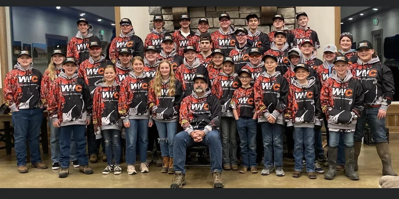 Group of children and an adult in matching black and red WIC sports jackets, posed indoors for a team photo in front of a stone fireplace, some children wearing baseball caps and others with hats or hoods, all smiling.