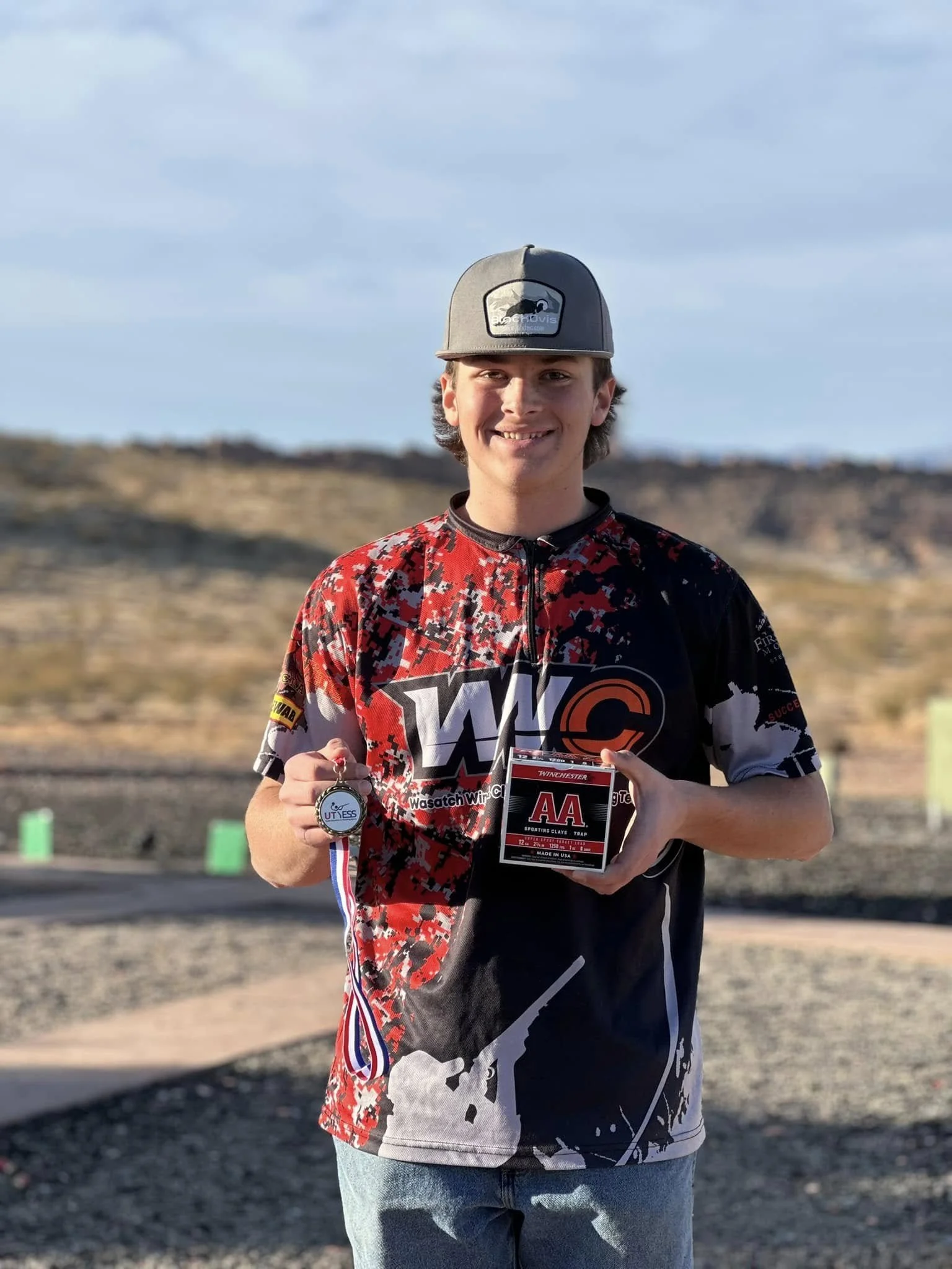 A young man wearing a black, red, and white jersey holding a box and a silver medal at an outdoor shooting range, smiling at the camera.