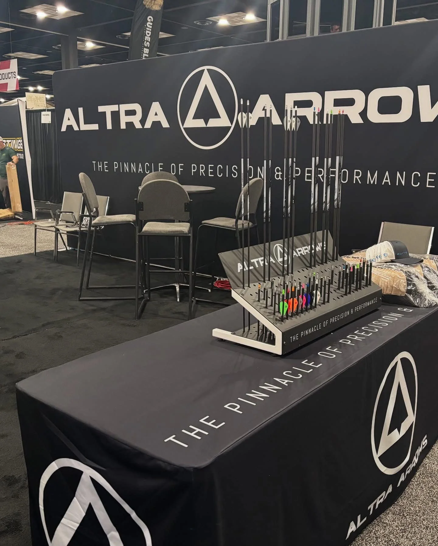 A display booth for Altra Arrow showcasing archery equipment, with a table covered in a black tablecloth featuring the company's logo and slogan, and a stand with multiple colorful arrows.
