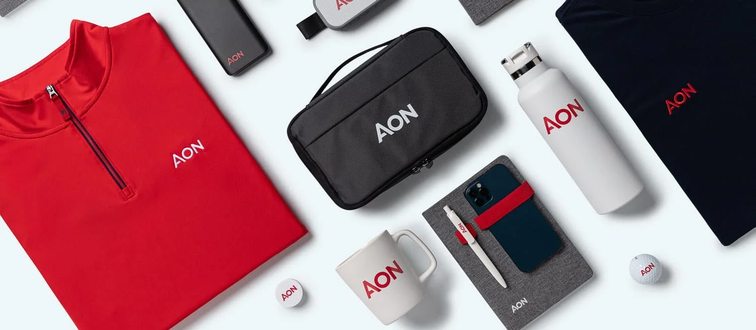 A flat lay of various branded items including a red quarter-zip fleece, a black pencil case, a white water bottle, a gray notebook with a pen and phone holder, a mug, and several small electronic devices, all featuring the AON logo.