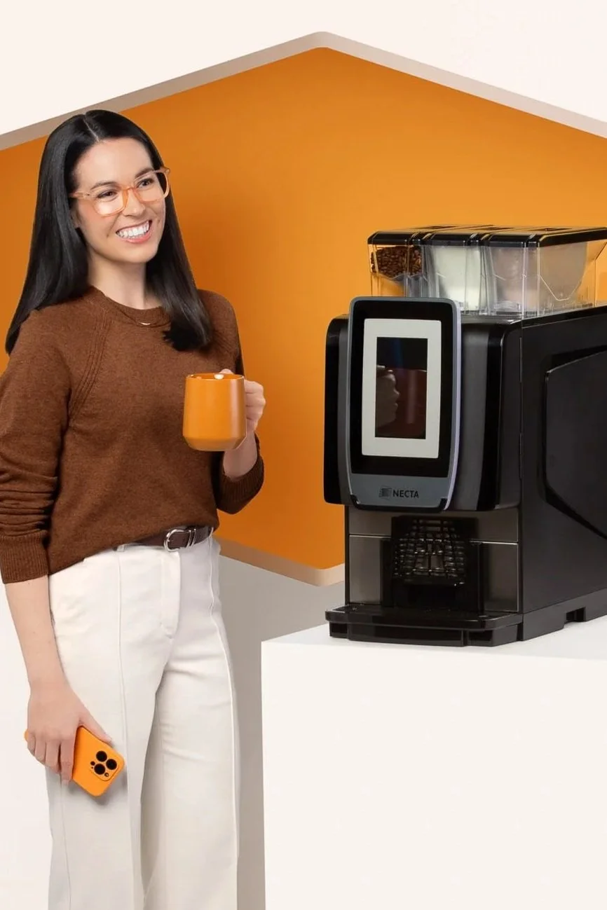 Woman with long dark hair, glasses, wearing a brown sweater and white pants, holding a coffee mug and smartphone, standing next to a coffee machine in a brightly colored room.