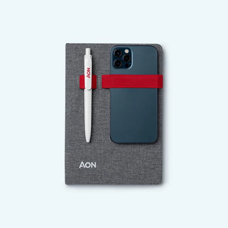 A gray AON notebook with a white AON pen attached by an elastic loop and a blue smartphone secured by a red elastic band, on a light blue background.