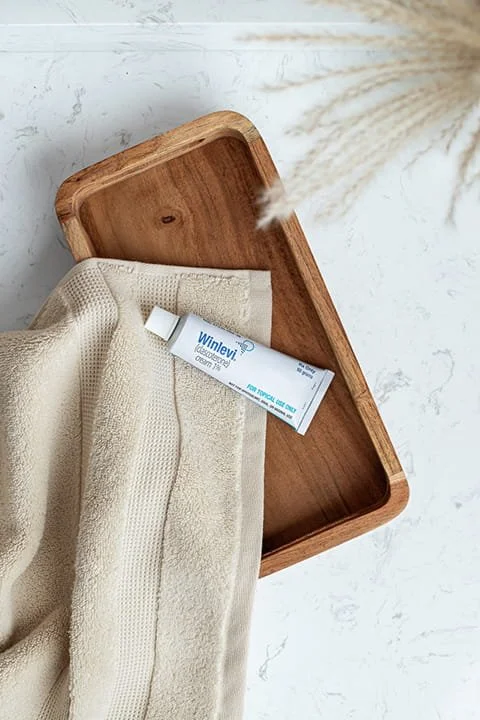 A wooden tray with a tube of hand cream and a cream-colored towel on a marble surface with a dried plant in the background.