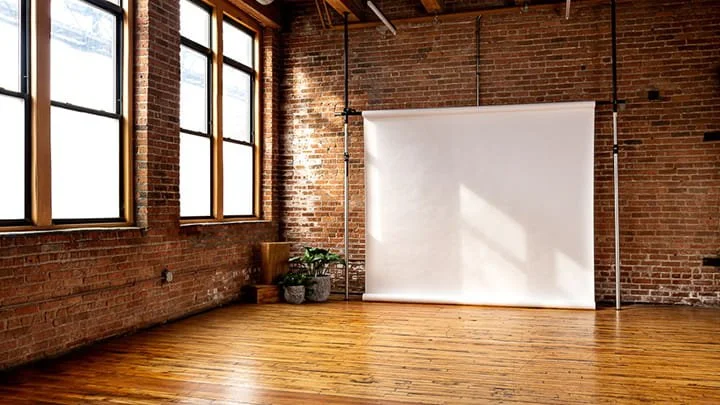 Empty photography or studio space with brick walls, wooden floors, large windows, and a white backdrop.