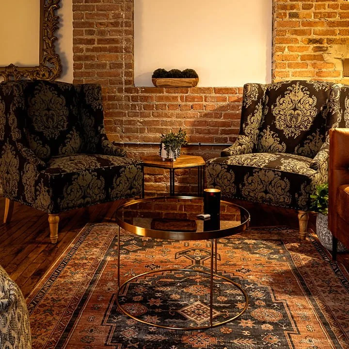 Living room with two patterned armchairs, a round glass coffee table with a candle and remote, a vintage rug, a brick wall with a small shelf, a mirror, and a potted plant.