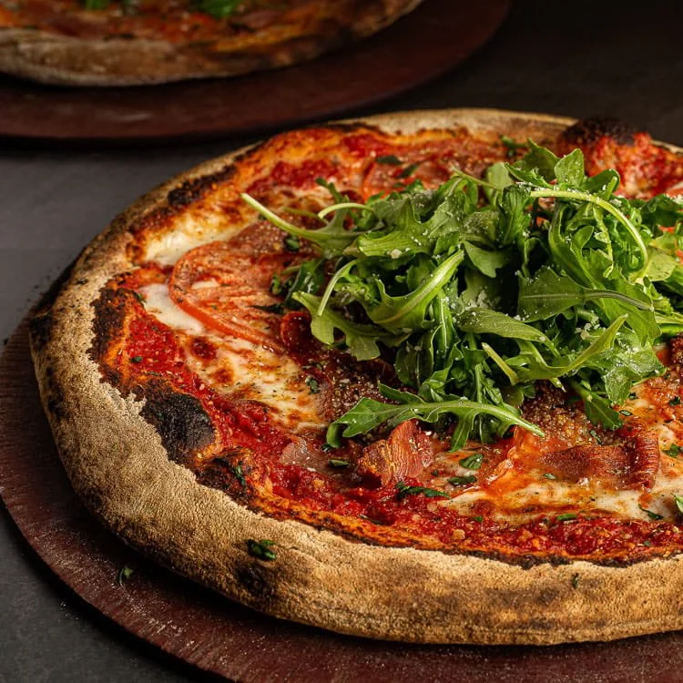 A close-up of a pizza with a charred crust, topped with tomato sauce, melted cheese, slices of pepperoni, bacon bits, and a handful of fresh arugula in the center.