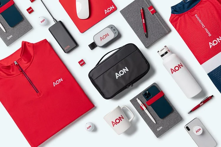 A flat lay of branded AON promotional items including a red quarter-zip jacket, water bottle, USB drives, a Wi-Fi hotspot device, notepad, pens, a mug, a smartphone, and a small bag, displayed on a light-colored surface.