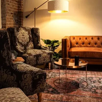 Living room with patterned armchairs, a brown leather tufted sofa, a round glass coffee table, a potted plant, and a floor lamp with a yellow shade.