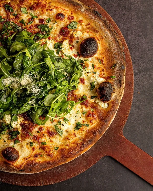 Close-up of a pizza with cheese, black olives, and fresh green arugula on top, served on a wooden pizza paddle.