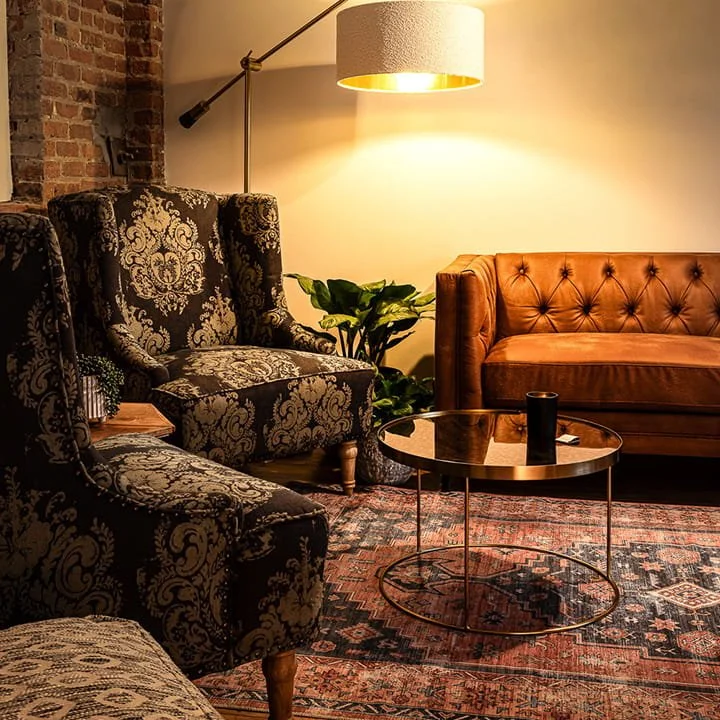 Cozy living room with vintage patterned armchairs, a leather sofa, a round glass coffee table, a large wall-mounted lamp, a lush green plant, and a patterned rug.