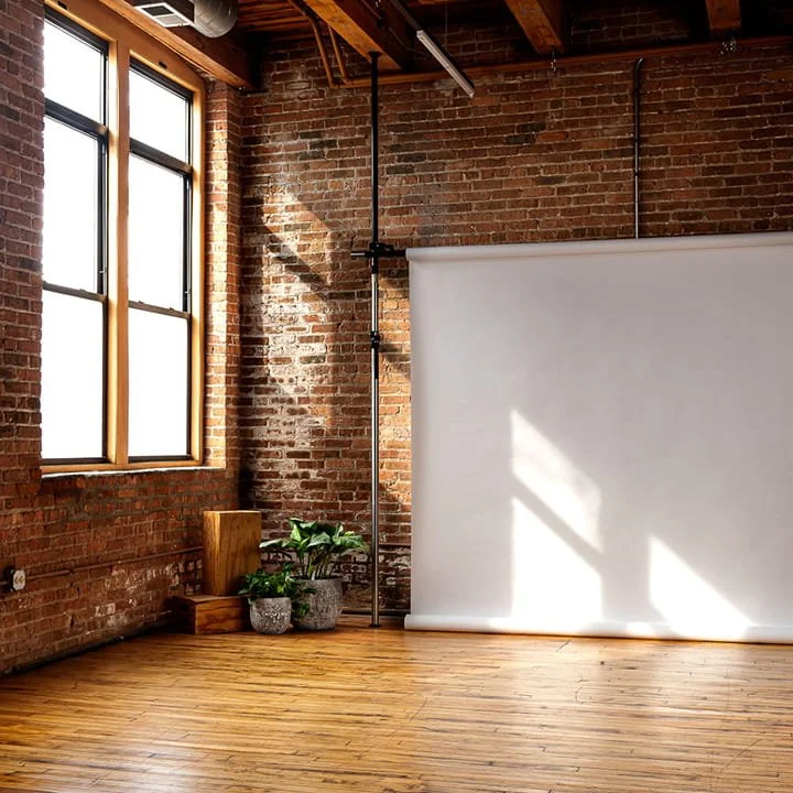 Empty room with brick walls, large windows, wooden floor, and a white backdrop roll, illuminated by natural light.