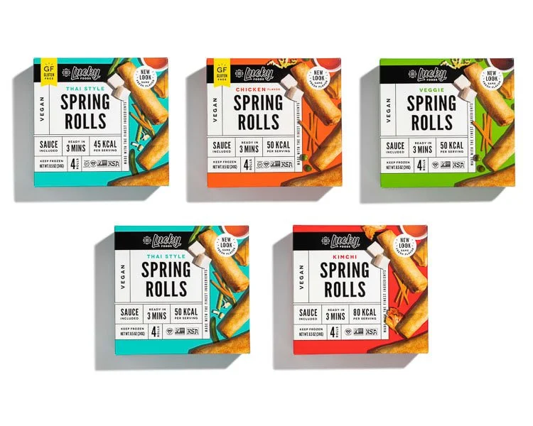 Five boxes of vegan spring rolls in various flavors, including Thai style, chicken, veggie, and kimchi, arranged in a row on a white surface.