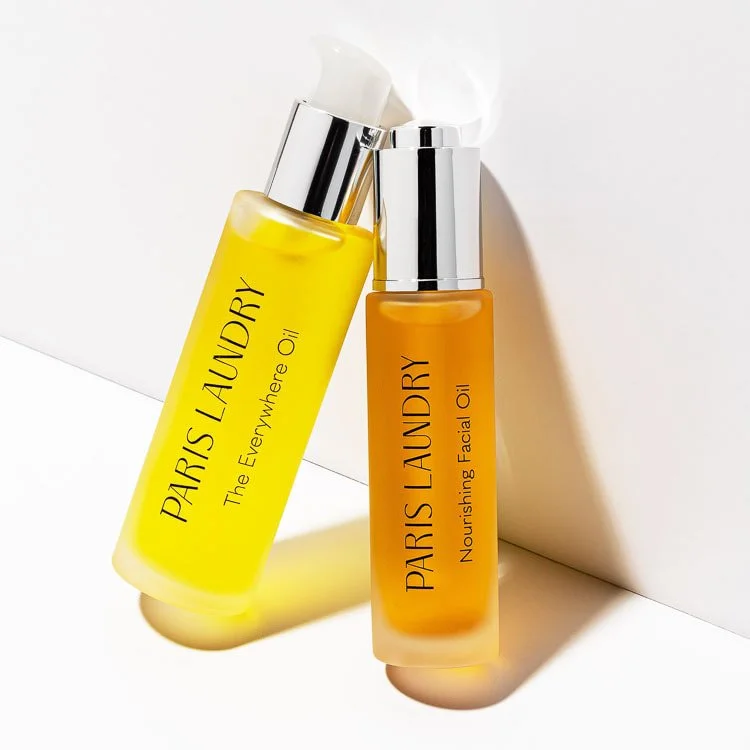 Two bottles of facial oil from Paris Laundry, one labeled 'The Everywhere Oil' in yellow and the other labeled 'Nourishing Facial Oil' in orange, with silver caps, resting on a white surface with shadows.