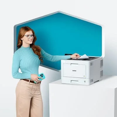 A woman with red hair and glasses holding a smartphone, standing next to a multifunctional printer in front of a blue background.