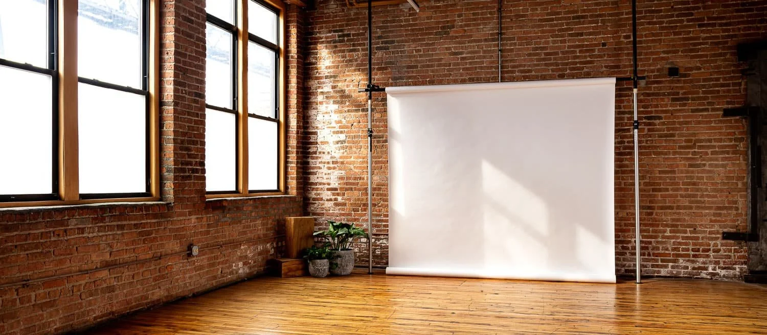 Empty photo studio with brick walls, wooden floor, large windows, and a blank white backdrop.