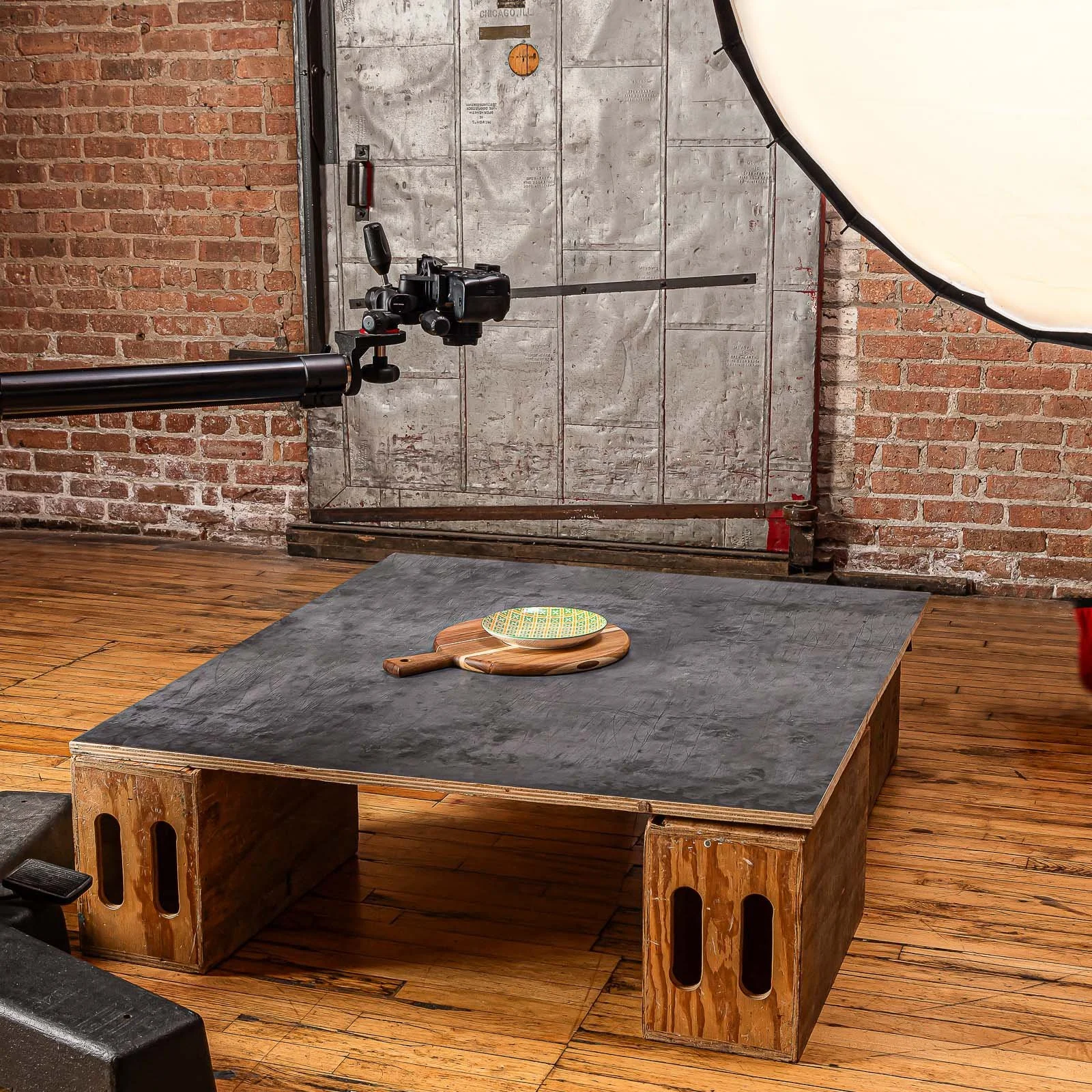A food photography studio setup with a camera shooting at a flat black table that has wooden cutting boards and a patterned bowl on top. The background features exposed brick walls, a metal door, and a large softbox light to the right.