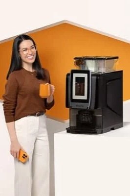 A woman standing next to a coffee machine, holding an orange mug in one hand and a phone in the other, in front of a white wall with an orange accent wall.