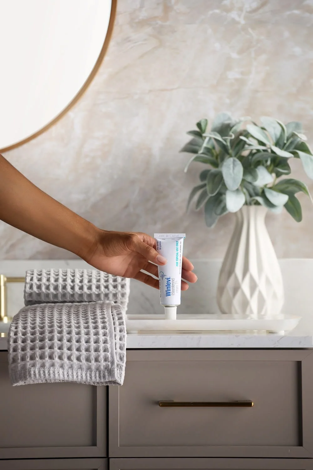 A hand holding a tube of Wenley dental cream over a bathroom sink, with a grey towel folded on the counter and a white vase with green foliage in the background.