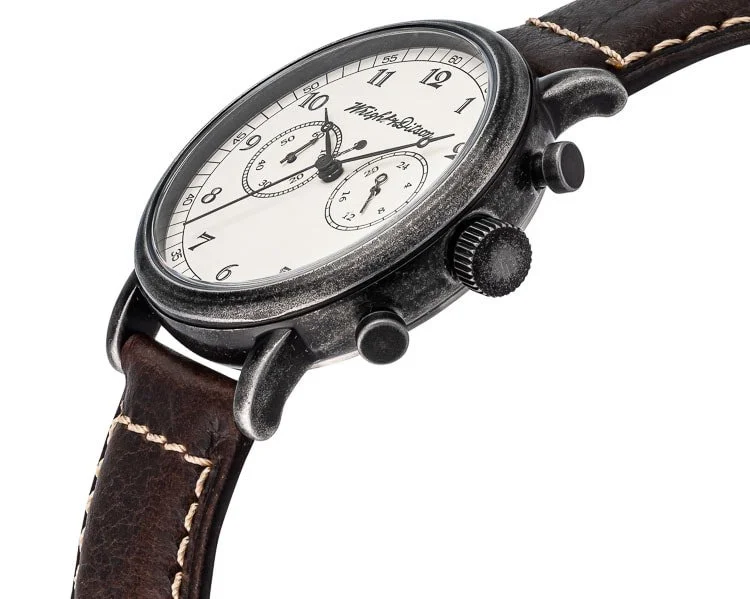 A wristwatch with a black metal case, white dial, black hands, and a brown leather band with white stitching.