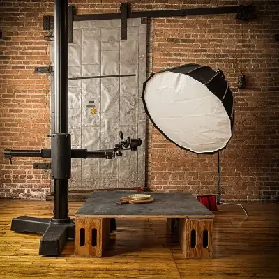 Photo of a photography studio setup with a black metal stand, a white umbrella reflector, and a wooden platform against exposed brick walls.