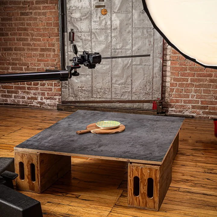 A photography setup with a camera mounted on a boom arm, professional lighting equipment, and a wooden platform with a gray top. A badminton racket and shuttlecock rest on the platform, set against a brick wall and a concrete door in the background.