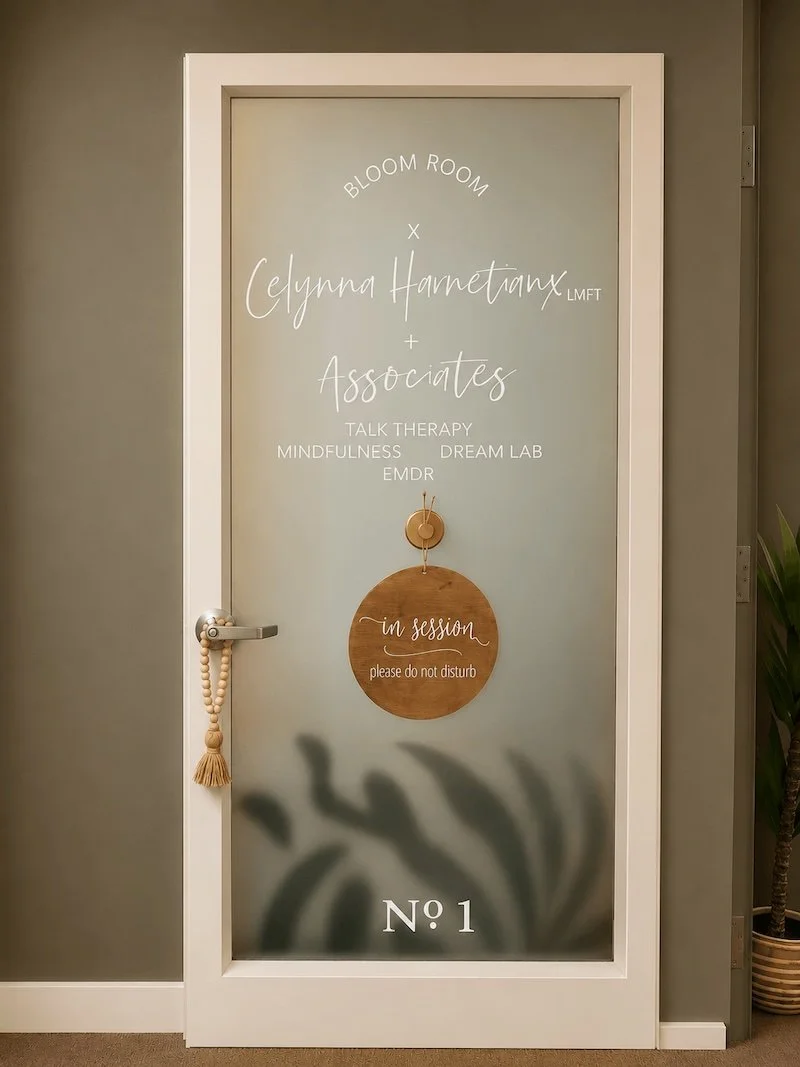 A door with a frosted glass panel that has the text 'Bloom Room, Celynn Harnetanyx LMFT & Associates' along with services like talk therapy, mindfulness, dream lab, and EMDR. A wooden sign hanging says 'in session, please do not disturb'. There is a door handle with a beaded tassel hanging from it, and the door has a plant silhouette at the bottom with the number 'Nº 1' at the base.