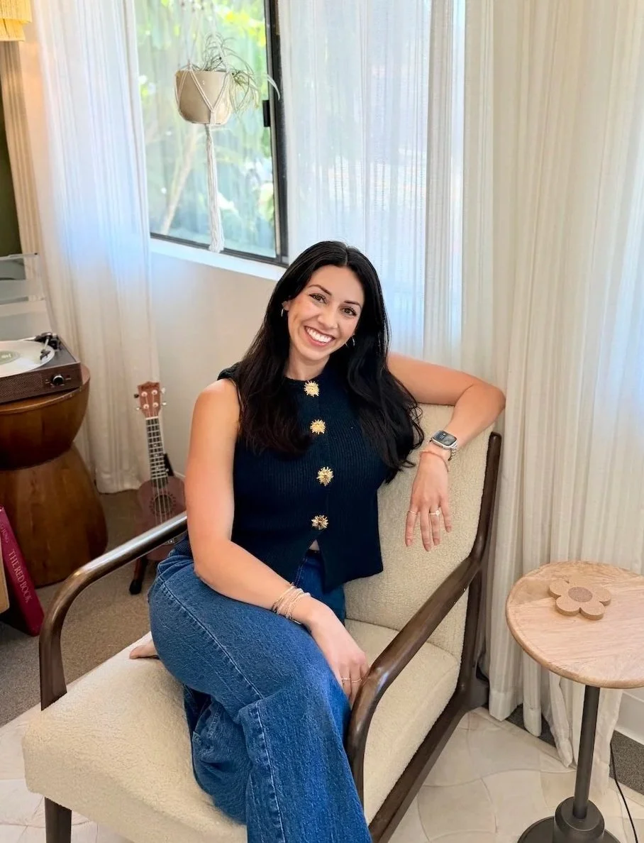 A woman with long dark hair smiling, sitting on a beige chair in a room with light-colored curtains and window. She is wearing a sleeveless black top with gold buttons and blue jeans, with a watch on her left wrist and bracelets on her right arm. There is a small round wooden table with a flower-shaped decor in the foreground, and a ukulele and a small record player in the background.