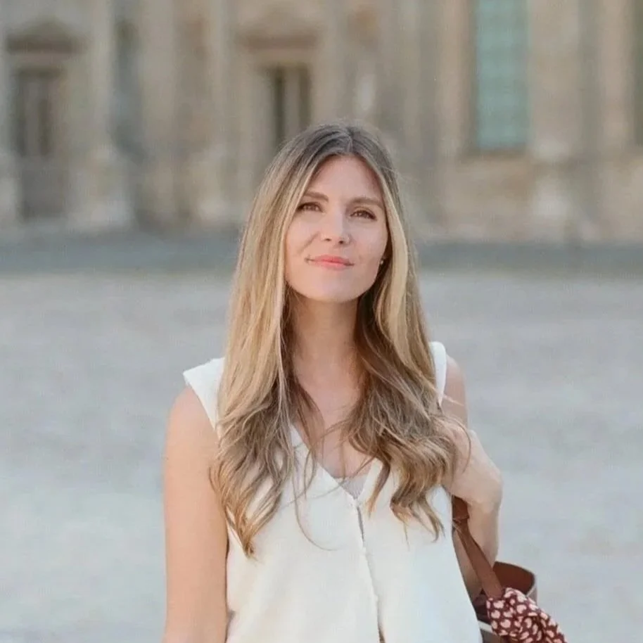 A woman with long blonde hair wearing a white sleeveless top and light-colored pants, standing outdoors in front of a historic building.