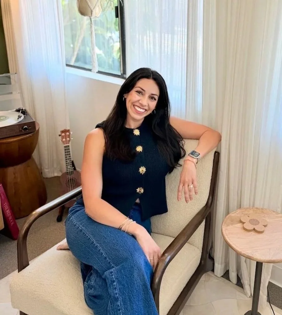 A woman with long dark hair smiling while sitting on a beige chair in a room with wooden and fabric furnishings, a small wooden side table with a flower-shaped decoration, a window with curtains, and a guitar in the background.