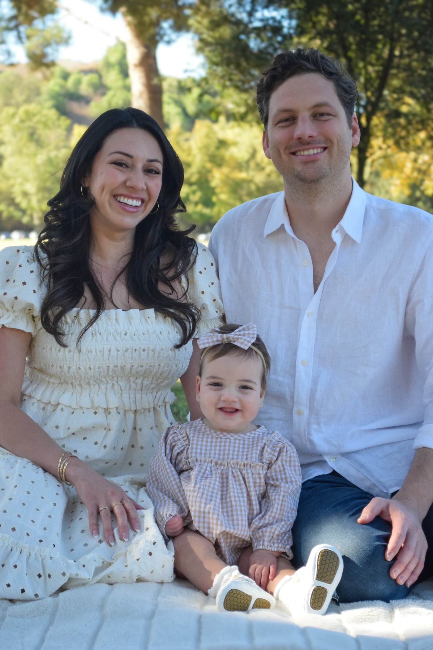 A smiling woman in a cream-colored dress with small holes, sitting outdoors with a man and a little girl. The woman has long, dark hair and wears gold jewelry. The man, with dark wavy hair, is wearing a white shirt. The little girl, sitting on the woman's lap, has light brown hair with a bow and is dressed in a checkered outfit. They are surrounded by green trees and sunlight.