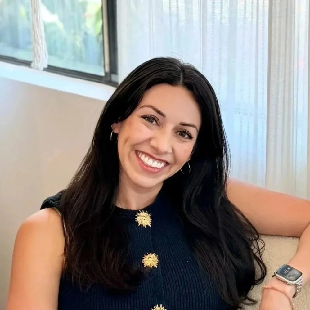 A woman with long black hair smiling, wearing a dark sleeveless top with decorative gold buttons, seated indoors with a window behind her.
