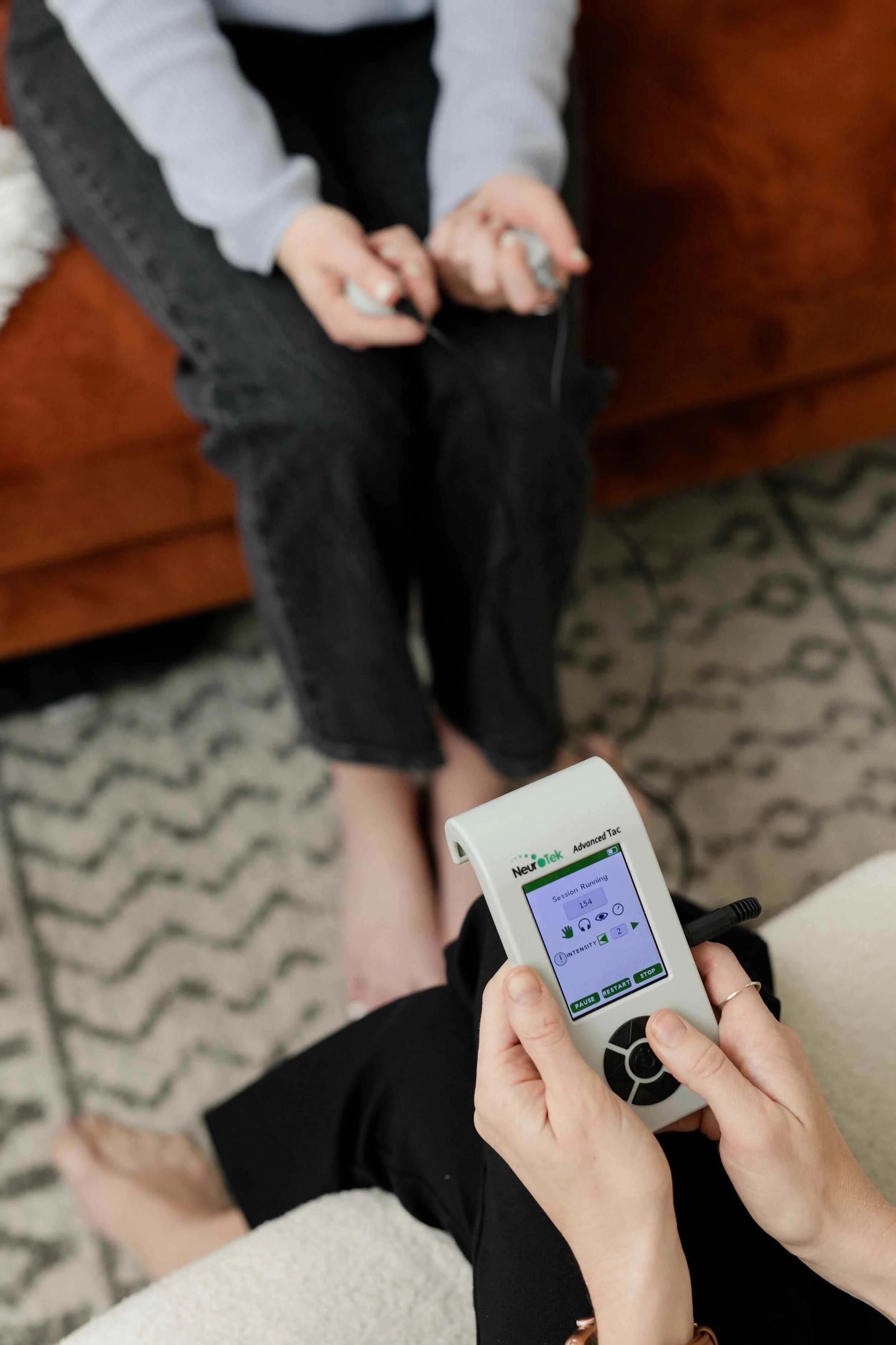 Person sitting on a chair with barefoot, holding a handheld device displaying settings, with another person resting their feet on this person's lap. Device is for bilateral stimulation for EMDR Therapy in South Orange County.