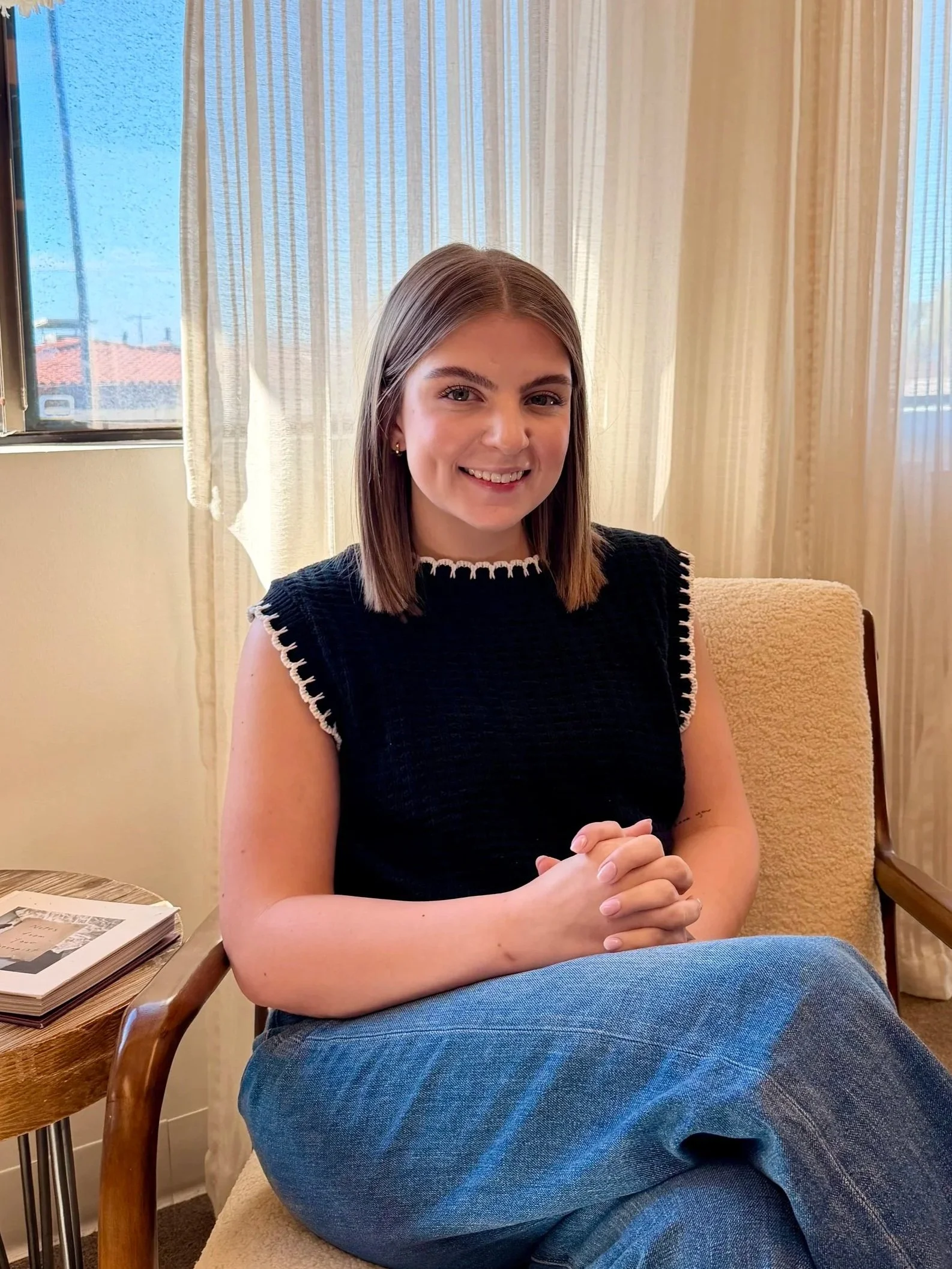 A young woman with wavy blonde hair smiling outdoors in a white dress with eyelet details, standing in front of greenery with sunlight.
