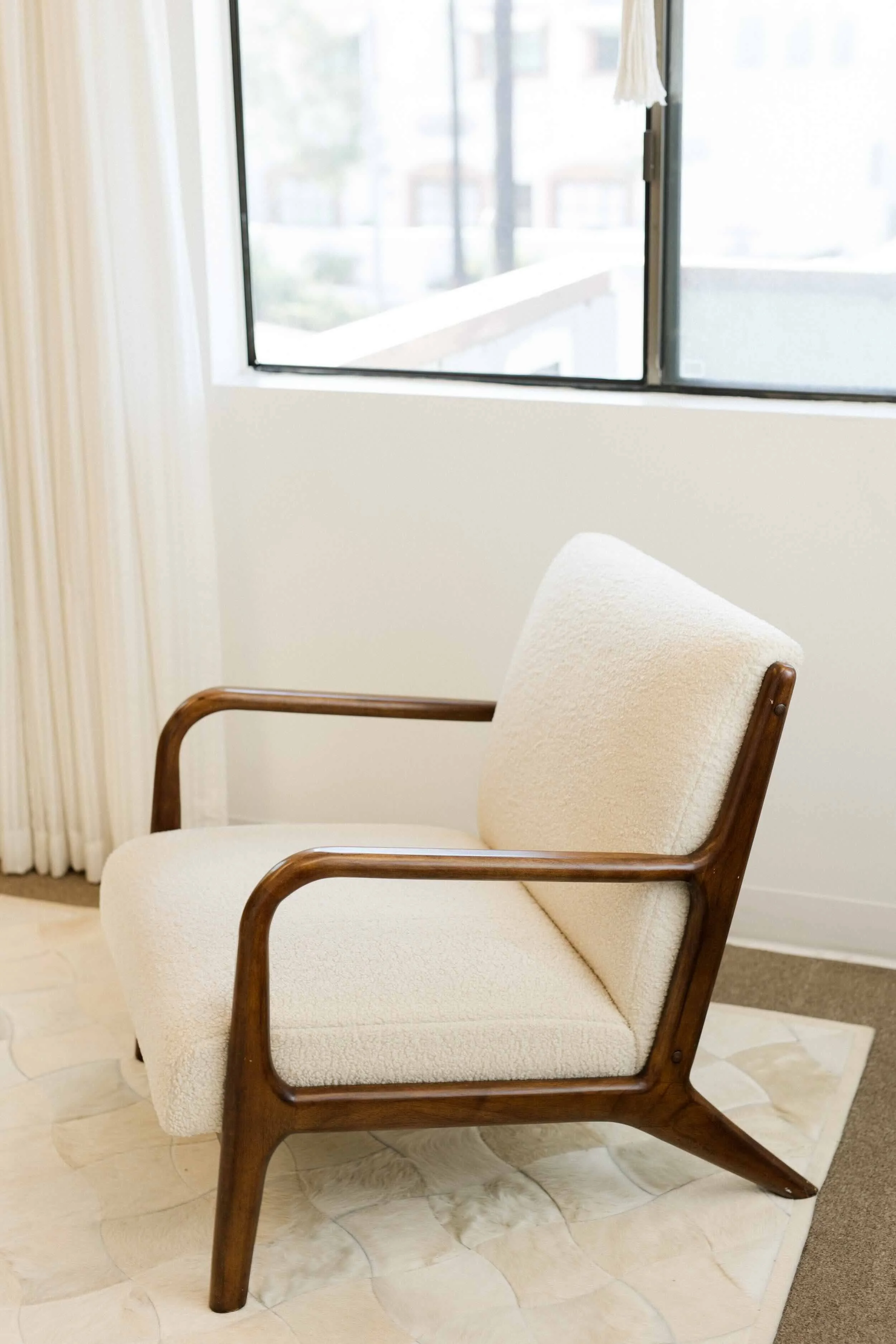A vintage-style armchair with wooden armrests and legs, upholstered in white fabric, placed on a tiled floor near a window with white curtains. Inside orange county, ca therapy office.
