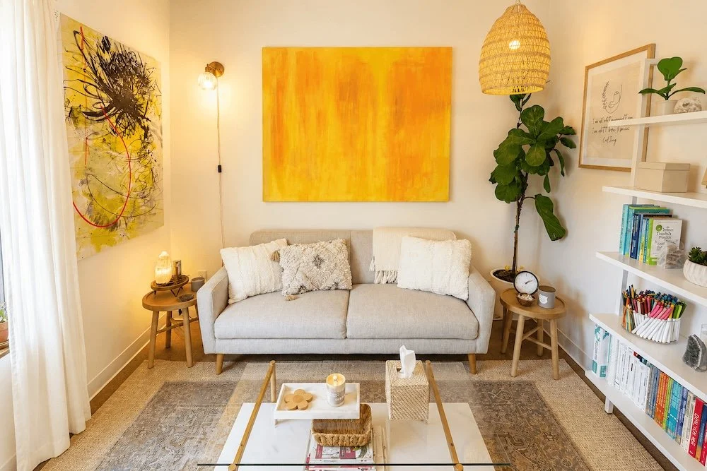 A cozy living room with a light beige sofa, decorated with white and grey pillows, against a white wall. Above the sofa hangs an abstract yellow painting, with a smaller abstract black and yellow piece on the adjacent wall. To the right of the sofa is a tall potted plant, and a round wooden side table with a clock and a candle. On the left side, there is a small wooden side table with a lamp and some decorations. A white rug covers the hardwood floor, and a coffee table with decorative items and a tissue box is in front. To the right, a white bookshelf contains books, plants, and decorative objects. A woven pendant lamp hangs from the ceiling.