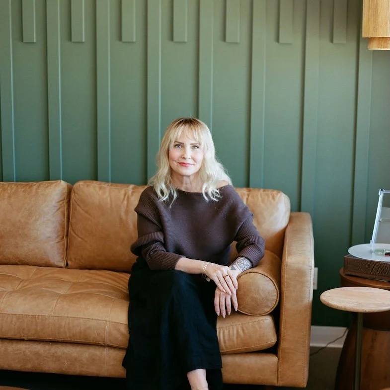 A woman with blonde, wavy hair and blue eyes holding a white coffee cup in a coffee shop, smiling at the camera.