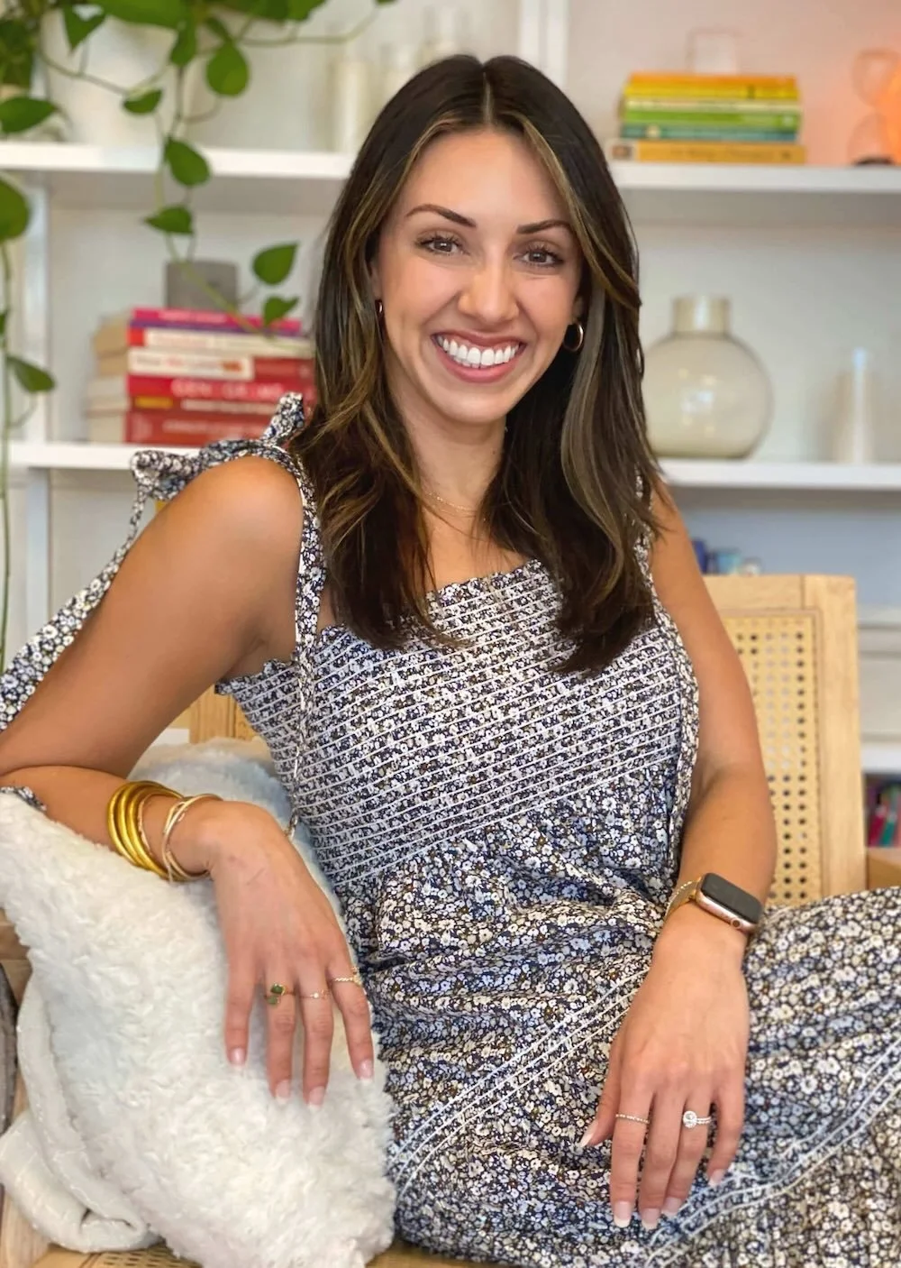 A woman with shoulder-length brown hair, smiling, sitting on a chair with a fluffy white pillow. She is wearing a patterned dress with shoulder bows and gold jewelry. There are books, a plant, and decorative items in the background.