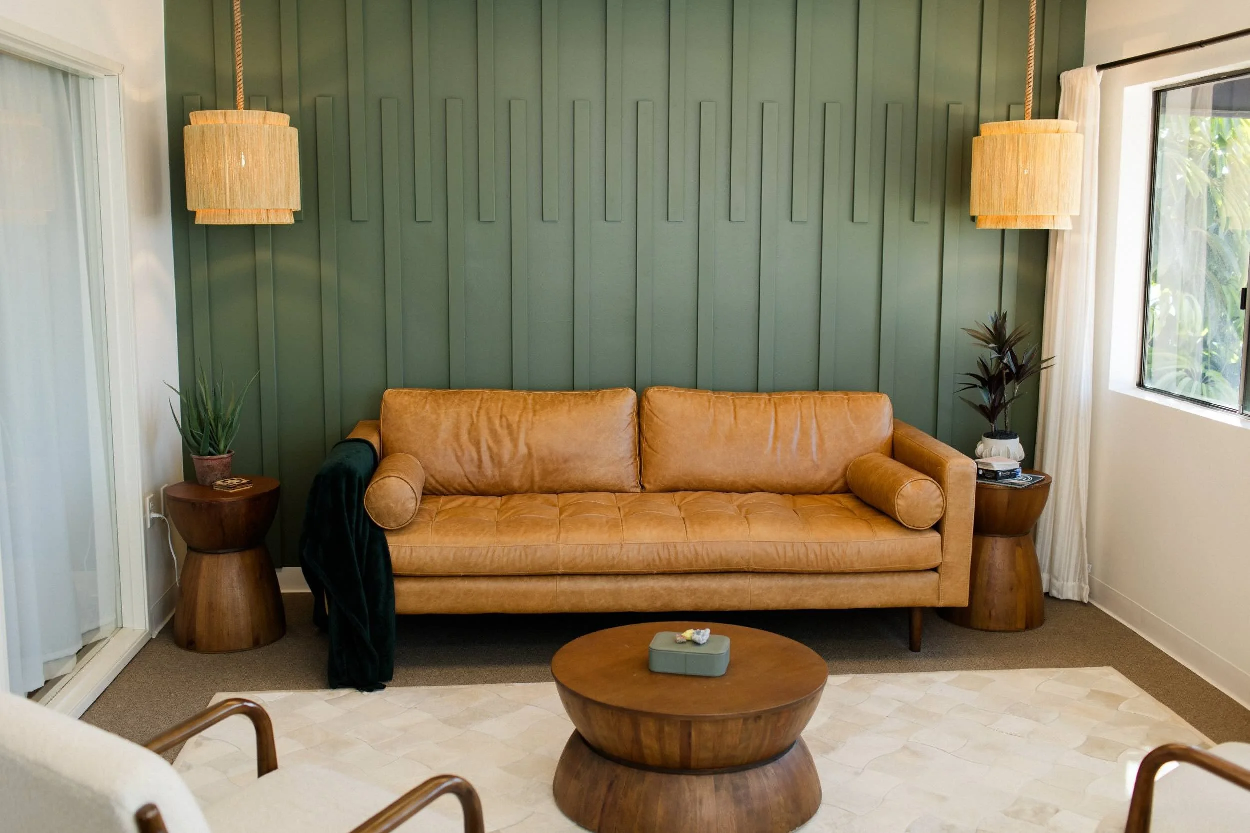 Living room with a brown leather sofa, two matching wooden side tables with potted plants, a round wooden coffee table, and a green paneled accent wall with three hanging tan fabric lamps. There's a window with white curtains on the right.
