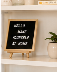 A black chalkboard with a wooden frame displaying the message 'HELLO MAKE YOURSELF AT HOME' in white letters, placed on a white shelf next to a potted green plant.