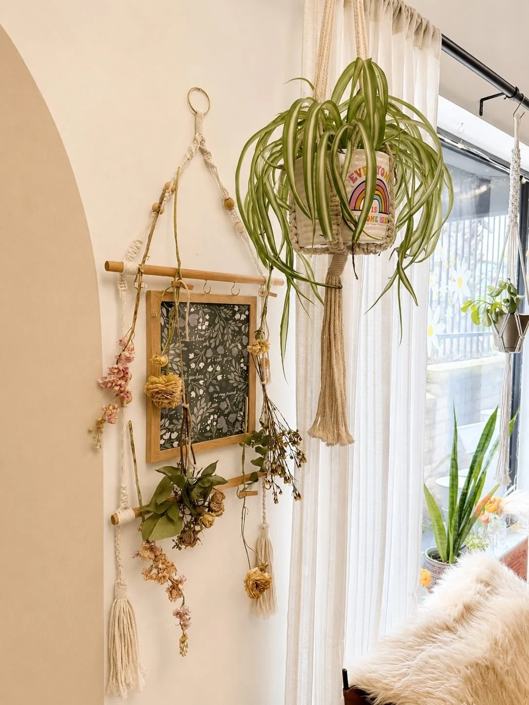 Indoor space with hanging plants, a decorative wall hanging with flowers and a chalkboard, near a window with sheer curtains and outdoor patio visible.
