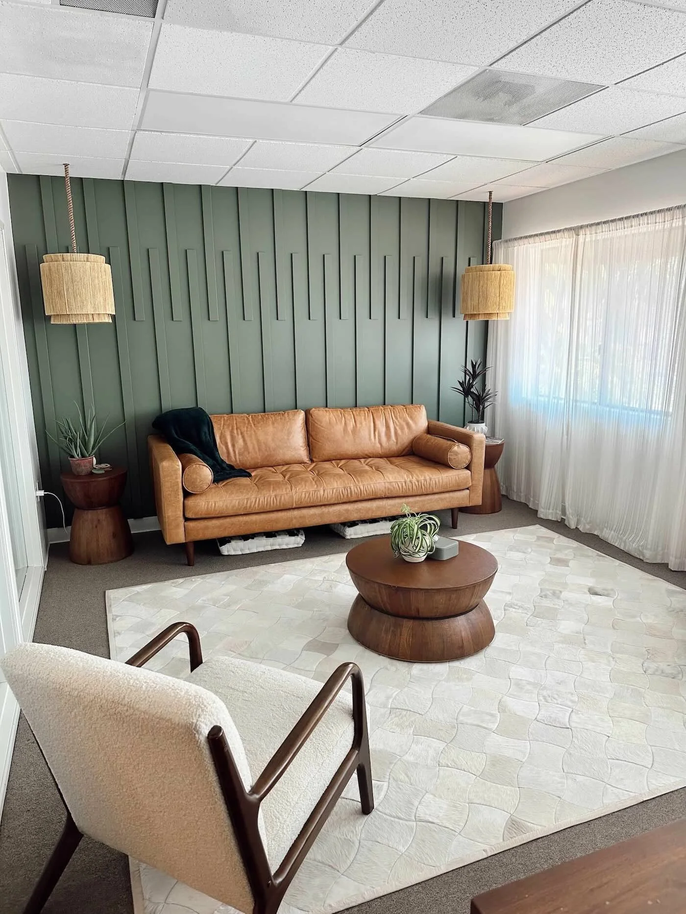 Living room with tan leather sofa, wooden coffee table, white area rug, beige armchair with wooden armrests, green plant, two beige hanging pendant lights, white sheer curtains, green accent wall with vertical paneling, ceiling with white tiles.
