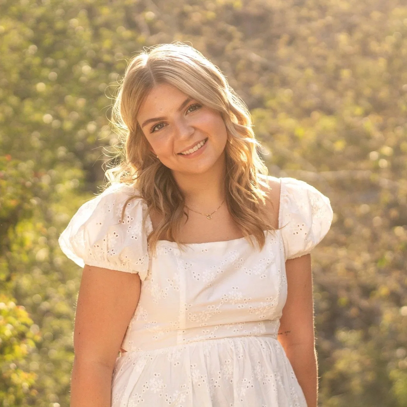 A young woman with blonde wavy hair smiling outdoors during golden hour, wearing a white eyelet dress with puffed sleeves.