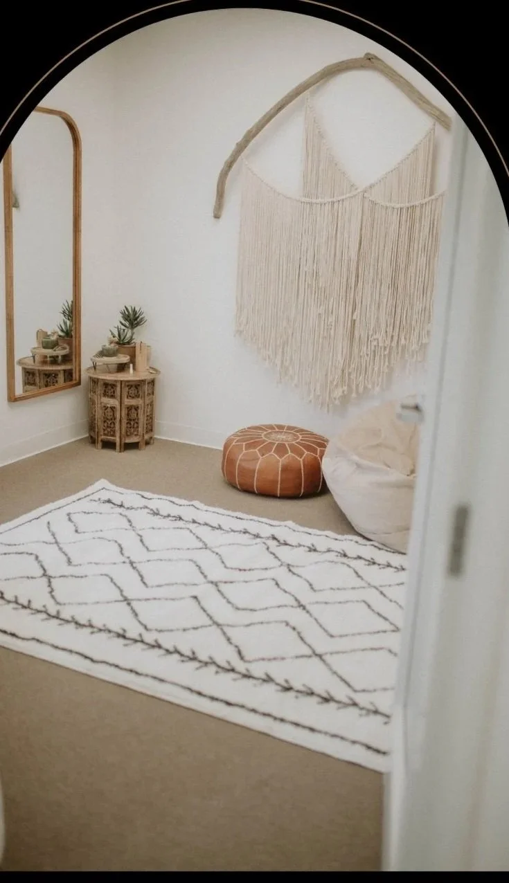 A cozy corner of a San Clemente therapy office with a boho-style wall hanging, a round leather pouf, a bean bag chair, a white patterned rug, a wooden mirror, and small plants on a wooden stand.