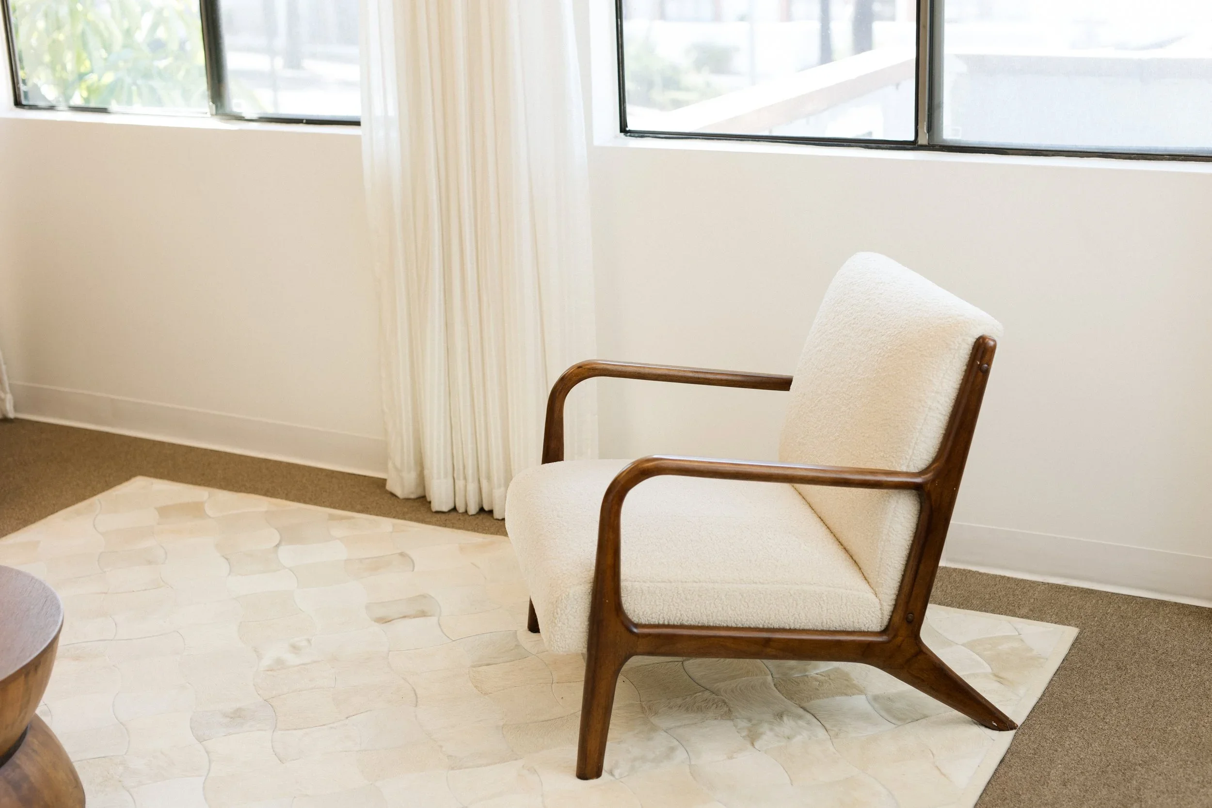 A beige armchair with wooden armrests next to a window with sheer white curtains. The room has light-colored walls, a patterned carpet, and a marble-like tiled floor.