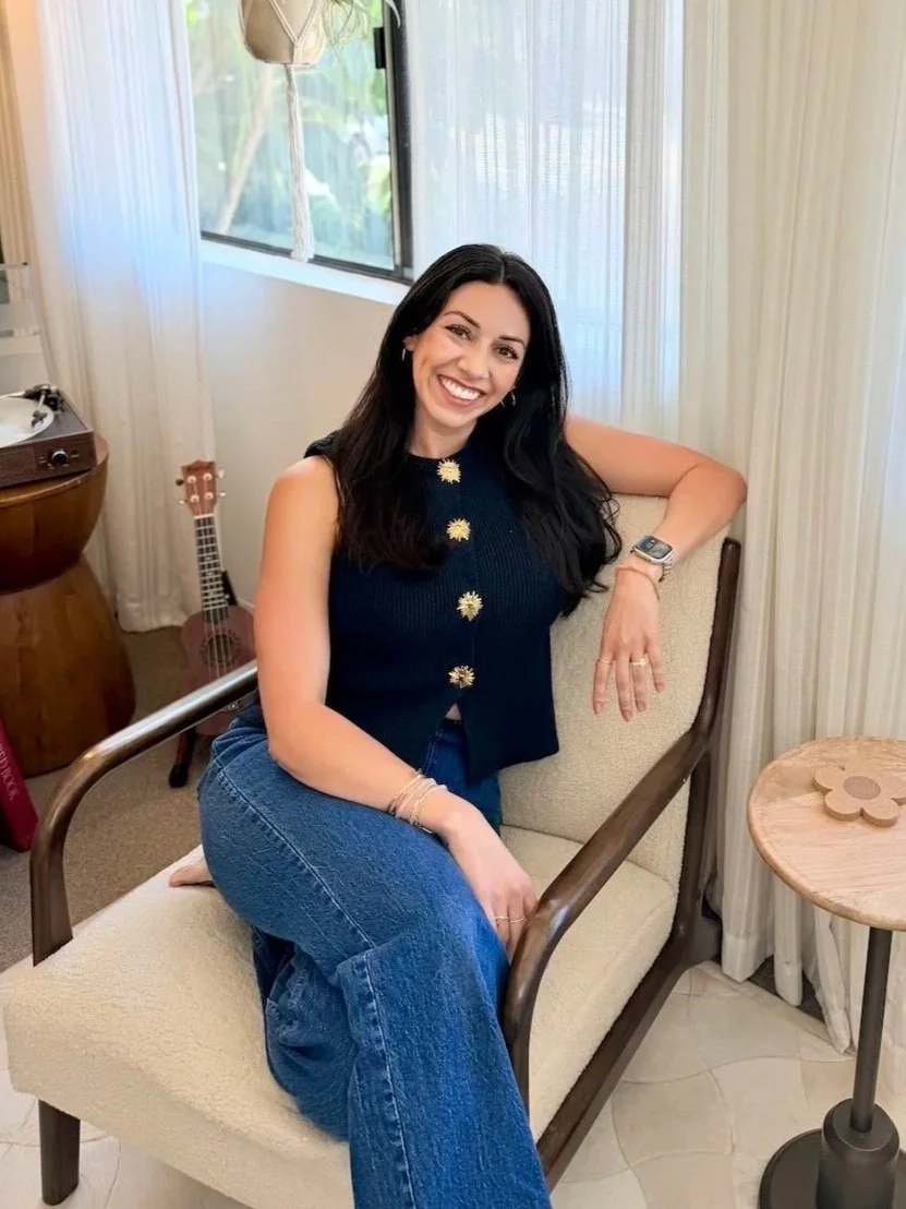 Smiling woman with dark hair sitting on a beige chair in a bright room with vertical blinds, a small wooden table with a flower-shaped decoration, a window showing greenery outside, a guitar, and a vintage record player in the background.