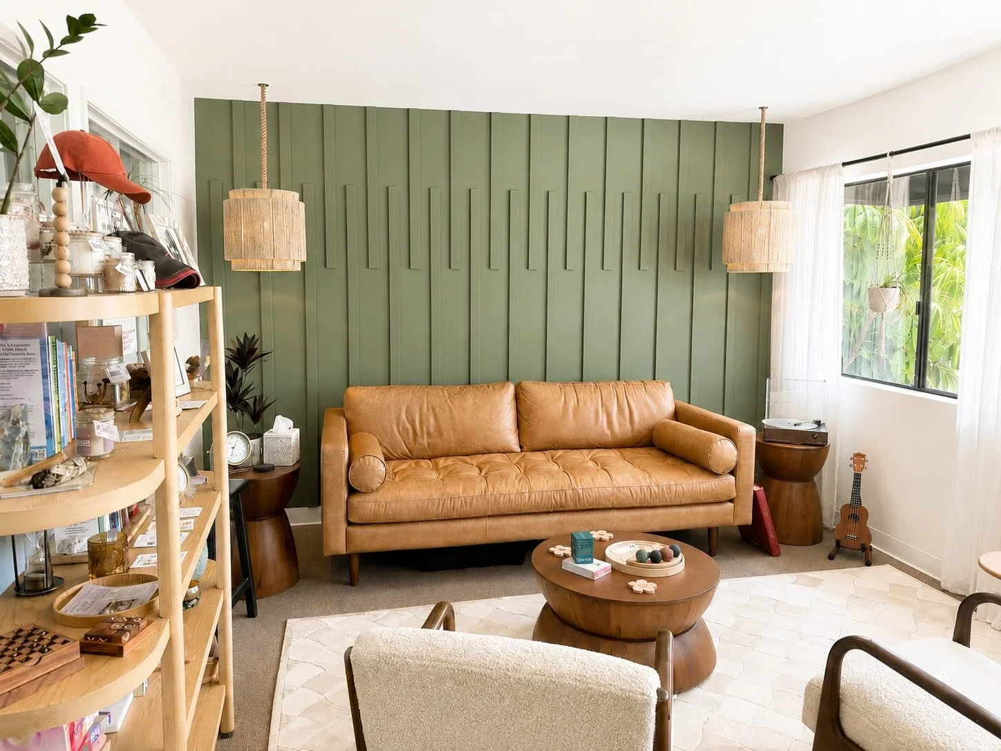 Living room with a tan leather sofa, green paneled accent wall, and wooden furniture, including a round coffee table and a bookshelf.