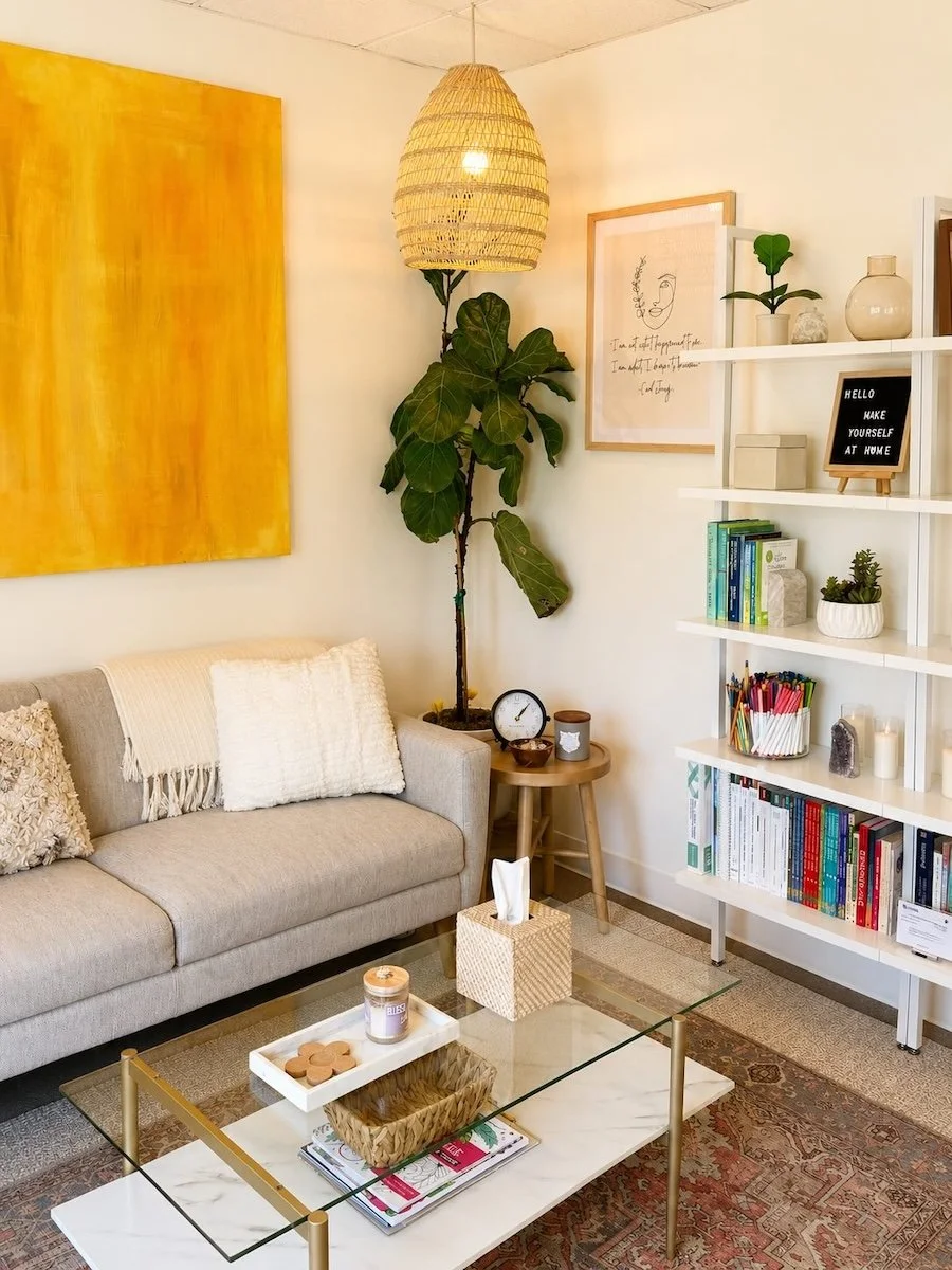 San Clemente therapy office with a beige sofa, white pillows, a glass coffee table with decor, a tall potted plant, a yellow wall art, a colorful bookshelf, and wall art
