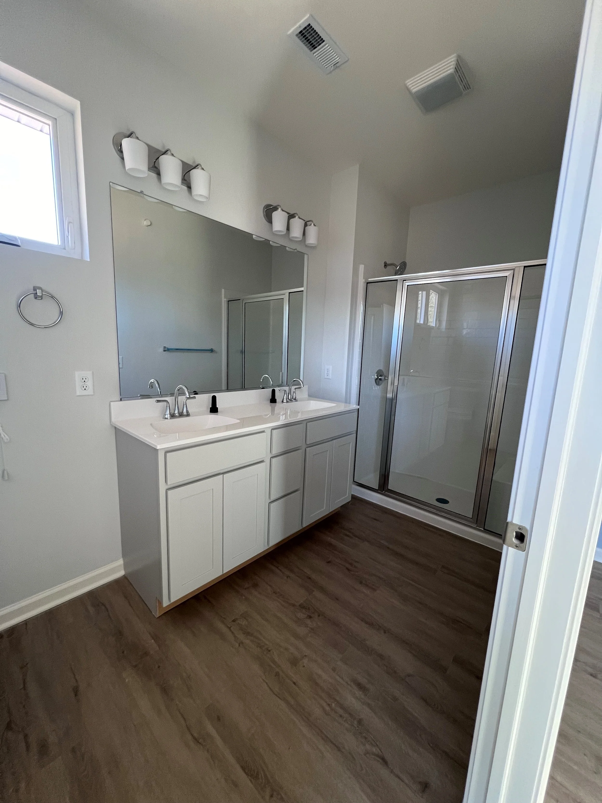 A modern bathroom with a double sink vanity, a large mirror, three-light fixture, separate shower with glass door, and a small window, with wood flooring.