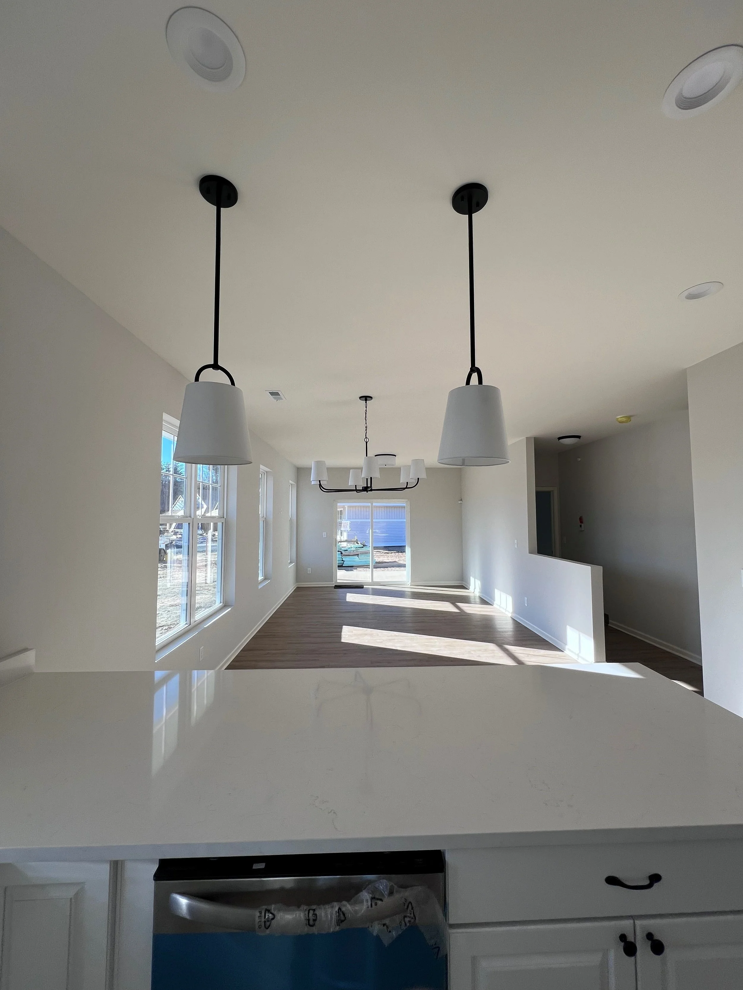 An empty modern dining and living area with beige walls, large windows, and hardwood floors. Multiple ceiling lights, a chandelier, and sliding glass doors are also visible.